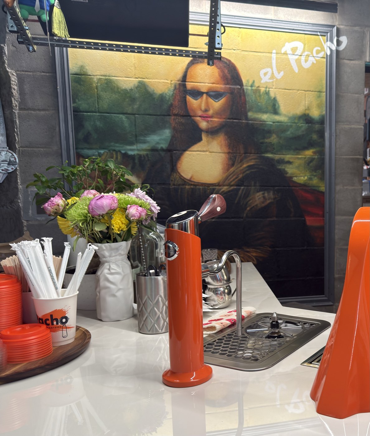 A Mona Lisa mural wearing sunglasses with el Pacho written beside it, above an orange beverage dispenser and a vase of flowers on the bar